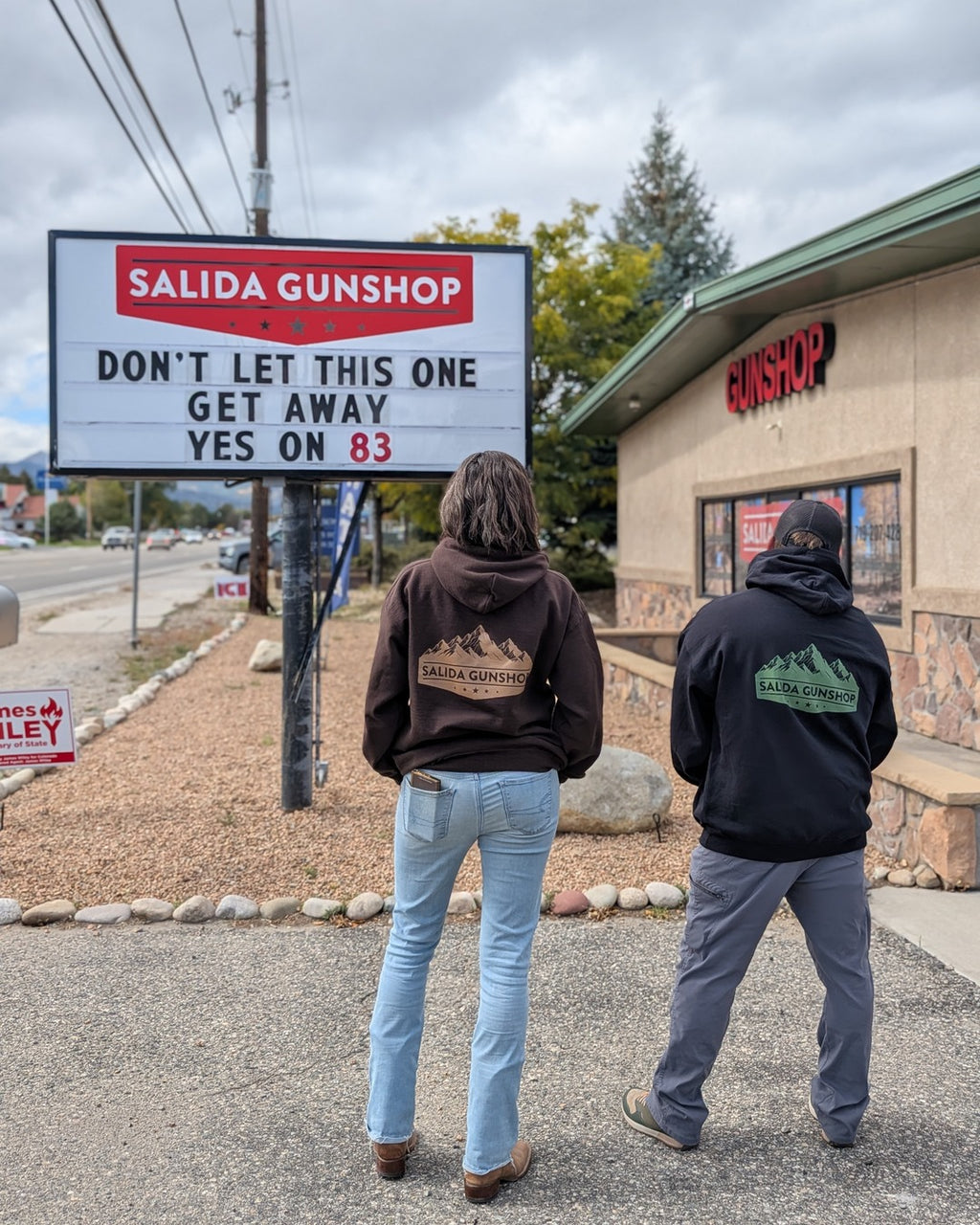 Two people wearing Salida Gunshop Mountain Logo Hoodies in front of the Salida Gunshop sign